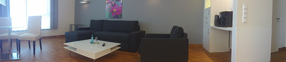 Wohnzimmer Dieses Bild zeigt das Wohnzimmer einer Familiensuite. In dem Zimmer stehen: ein Kaffeetisch mit einem Sessel und einem Sofa, ein kleiner Tisch mit zwei Stühlen und ein Regal, welches als Raumtrenner dient. Der Raum hat ein Großes Fenster.